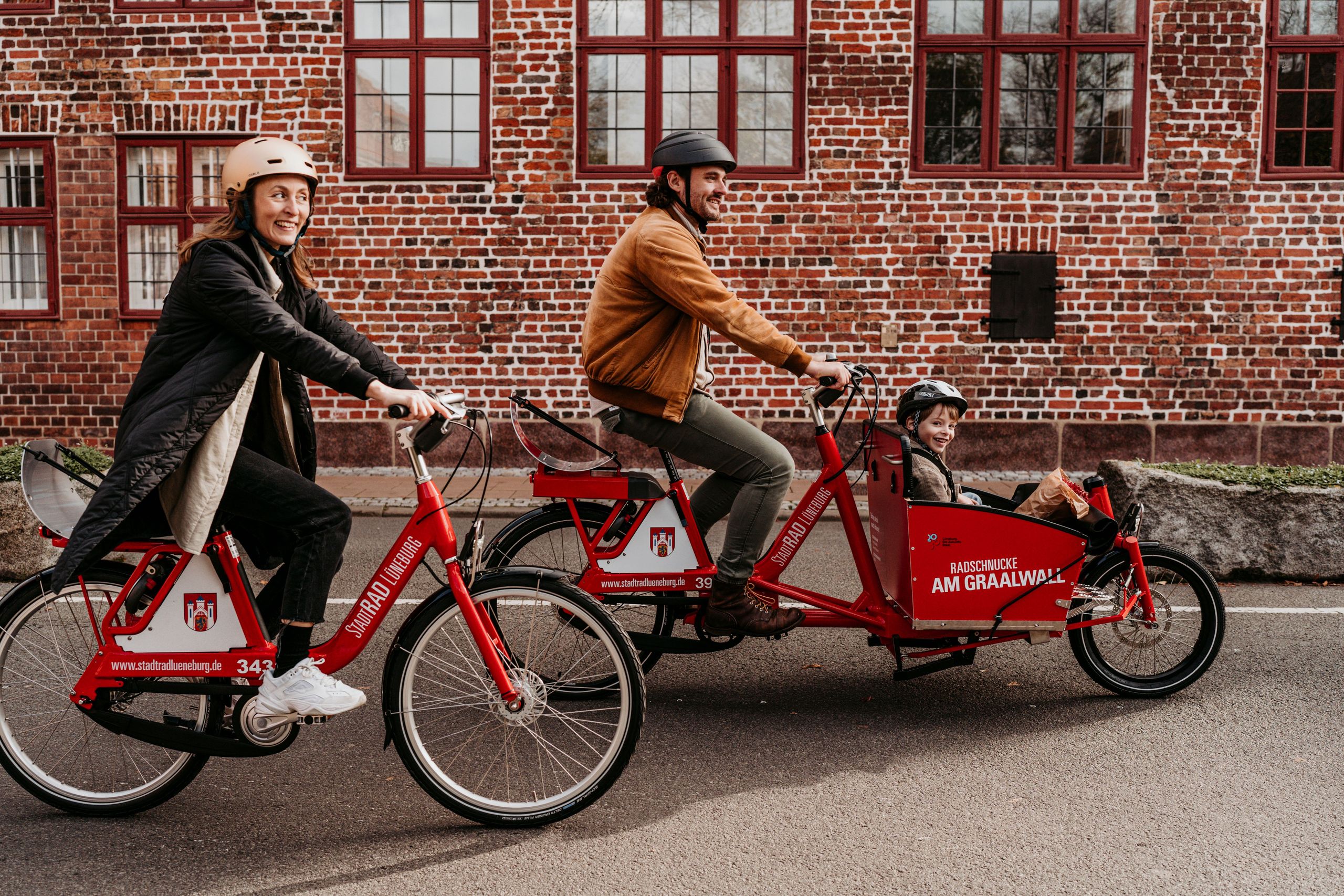 Eine Frau fährt auf einem Fahrrad und ein Mann fährt mit einem Lastenrad. Ein Kind sitzt im Korb des Lastenrads.